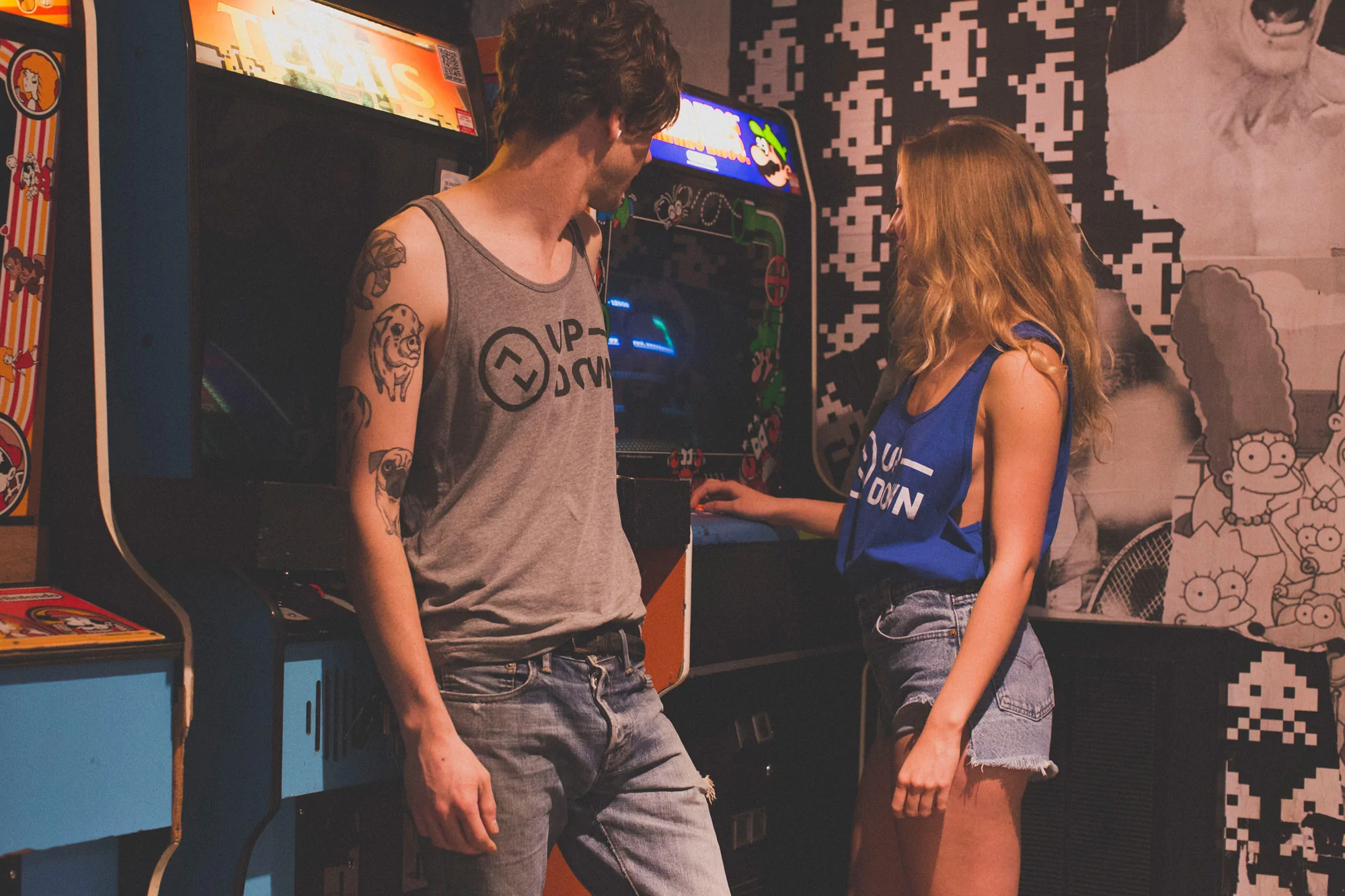 A young man and woman play retro arcade games in a vibrant gaming space, surrounded by colorful machines and playful decor.